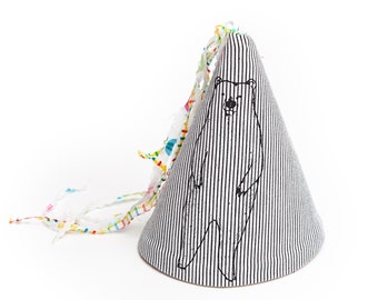 Pode incluir: Um chapéu de festa listrado branco e preto com uma ilustração em preto e branco de um urso. O chapéu tem uma guarnição colorida e franjada.