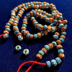 May include: Strands of handmade beads in shades of blue, brown, and white, strung on red thread. The beads have a speckled pattern. Two loose beads are also visible. The beads are displayed on a dark blue surface.