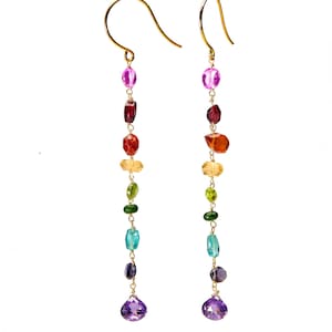 May include: Gold earrings with a cascade of colourful gemstones. The earrings feature a variety of shapes and hues, including purple, red, orange, yellow, green, blue, and a large purple teardrop at the bottom. The gemstones are strung on delicate gold chains.