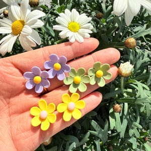Könnte beinhalten: Sechs Paare farbenfroher Ohrringe in Blumenform. Zwei Paare sind gelb, zwei grün und zwei lila. Jede Blume hat eine kleine gelbe Mitte.