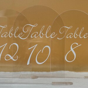 Puede incluir: Señales de números de mesa de acrílico transparente con arcos. Cada señal tiene la palabra "Table" en elegante escritura blanca, seguida de un número: 12, 10 y 8. Las señales se muestran sobre una superficie blanca contra un fondo dorado.