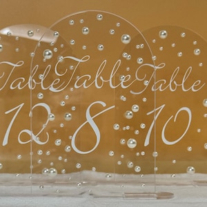 Puede incluir: Números de mesa de acrílico transparente con diseño de perlas y burbujas. Los números 12, 8 y 10 se muestran en escritura blanca, junto con la palabra "Table". Estos números de mesa son ideales para bodas y eventos.