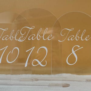 Puede incluir: Números de mesa de acrílico transparente con arcos. Cada letrero muestra la palabra "Table" en escritura elegante, seguida de los números 10, 12 y 8. Ideales para la decoración de mesas. Los letreros son de acrílico transparente.