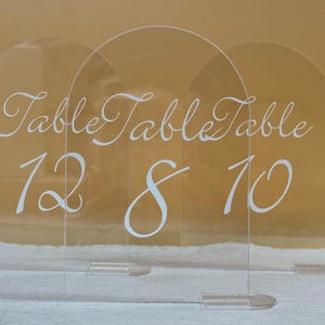 Puede incluir: Señales de número de mesa de acrílico transparente con arcos en la parte superior. El texto blanco en cursiva dice "Table" sobre los números 12, 8 y 10. Cada señal tiene una base transparente. Ideal para bodas y eventos.