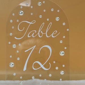 Puede incluir: Número de mesa de acrílico transparente con una parte superior arqueada, que muestra la palabra "Table" y el número "12" en escritura blanca. El letrero está adornado con adornos de perlas dispersas. Un pequeño soporte sostiene el letrero.
