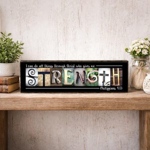 May include: A black wooden sign with the word "STRENGTH" spelled out in various textures and colors. The sign includes the text "I can do all things through Christ who gives me - Philippians 4:13."