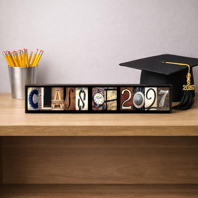Puede incluir: Un letrero rectangular de madera que muestra la palabra CLASS 2027, donde cada letra y n&uacute;mero est&aacute; formado por una fotograf&iacute;a de un objeto. El letrero se encuentra junto a un portal&aacute;pices plateado con l&aacute;pices amarillos y un birrete negro.