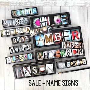 May include: A collection of personalized name signs in various sizes and styles. Each sign features individual letters with unique backgrounds, framed in dark wood. The signs spell out names like "Austin," "Wilson," and "Chelsea." The text "SALE - NAME SIGNS" is at the bottom.