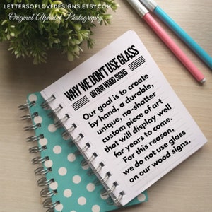 May include: A close-up of a white notebook with a black and white sign that reads "WHY WE DON'T USE GLASS ON OUR WOOD SIGNS". The sign explains that the goal is to create durable, unique, and custom pieces of art that will last for years to come. The notebook is on a wooden surface with a green plant in the background.