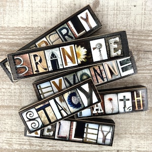 May include: Wooden name blocks with individual letters, each block spelling out a different name. The letters are set against various backgrounds, including floral, architectural, and natural elements. The blocks are in a dark brown frame.