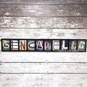 Puede incluir: Una obra de arte enmarcada que deletrea el nombre "GENCARLA" utilizando bloques de letras individuales. Cada letra presenta una imagen de fondo &uacute;nica, con un fondo de madera envejecida. El texto "Letters of Love Designs" est&aacute; en la parte inferior.