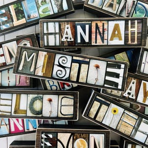 May include: Wooden name blocks with individual letters in various fonts and styles. Each block is framed in a dark wood. Some letters feature decorative elements like flowers and swirls. The name "Hannah" is visible.