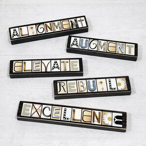 May include: Five rectangular wooden boxes with words spelled out in individual letters. The words are "ALIGNMENT", "AUGMENT", "ELEVATE", "REBUILD", and "EXCELLENCE". The letters are in various colors and styles, with decorative elements.