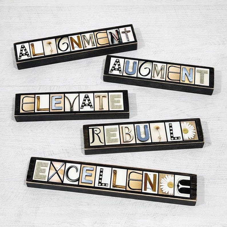 May include: Five rectangular wooden boxes with words spelled out in individual letters. The words are "ALIGNMENT", "AUGMENT", "ELEVATE", "REBUILD", and "EXCELLENCE". The letters are in various colors and styles, with decorative elements.