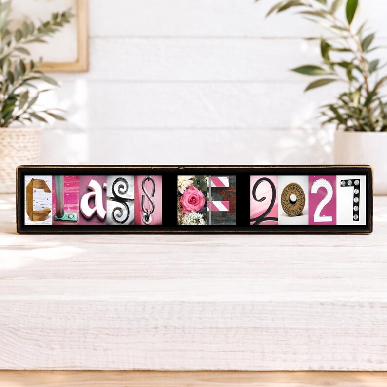 Puede incluir: Un marco negro rectangular que contiene una serie de azulejos fotogr&aacute;ficos cuadrados que deletrean la palabra CLASS 2027. Las letras y n&uacute;meros est&aacute;n formados por varios objetos, incluyendo una C de madera, una rosa rosa y una moneda de metal.