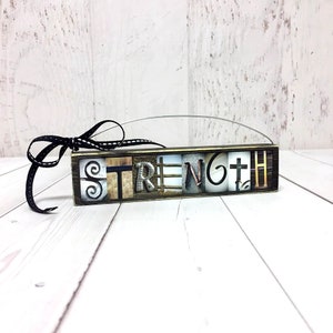 May include: A wooden sign with the word "STRENGTH" spelled out in mixed metal letters. The sign is hanging from a black ribbon with white polka dots.