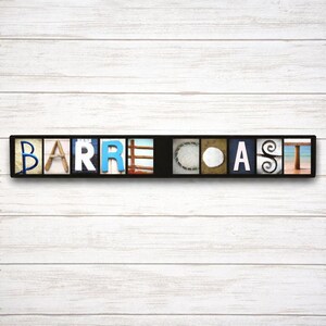 Puede incluir: Un letrero rectangular negro con la palabra "BARRE COAST" deletreada en bloques de letras individuales. Cada bloque de letras tiene un dise&ntilde;o &uacute;nico, incluyendo im&aacute;genes del oc&eacute;ano, conchas y madera.