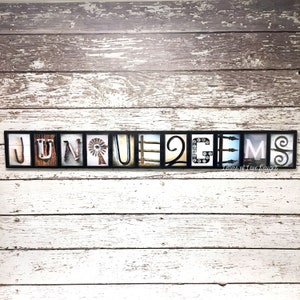 Puede incluir: Una pieza de arte mural enmarcada que deletrea "JUNE GEMS" en baldosas de letras individuales. Cada letra presenta un dise&ntilde;o &uacute;nico, que incluye un molino de viento, una valla y elementos decorativos. El fondo es una pared de madera blanca y gris desgastada.