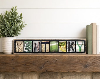 May include: A wooden sign spelling "Kentucky" with each letter featuring a different image. The letters are set in a black frame and rest on a wooden shelf. A potted plant and books are in the background.