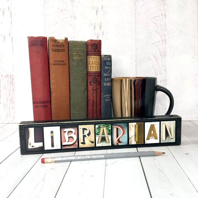 Puede incluir: Un letrero decorativo de madera con la palabra "LIBRARIAN" escrita con letras coloridas. El letrero est&aacute; frente a una fila de libros antiguos y una taza de caf&eacute;. Un l&aacute;piz con el texto "BETTER OF LOVE DESIGNS" est&aacute; frente al letrero.