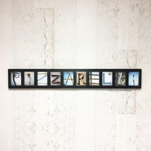 Puede incluir: Una obra de arte mural enmarcada en negro que deletrea el nombre "Pizzarelli" con baldosas de letras individuales. Cada baldosa presenta una imagen &uacute;nica, que incluye una flor, un velero y un faro. El fondo es una pared de madera blanca.