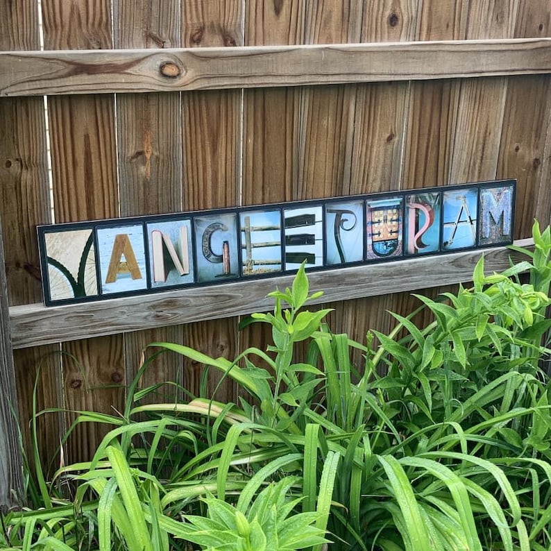 Puede incluir: Un letrero rectangular de madera con la palabra "MANGEETERIAM" escrita en bloques de letras individuales. Las letras son de varios colores y estilos, sobre un fondo azul y blanco. El letrero est&aacute; montado en una valla de madera, con plantas verdes en primer plano.