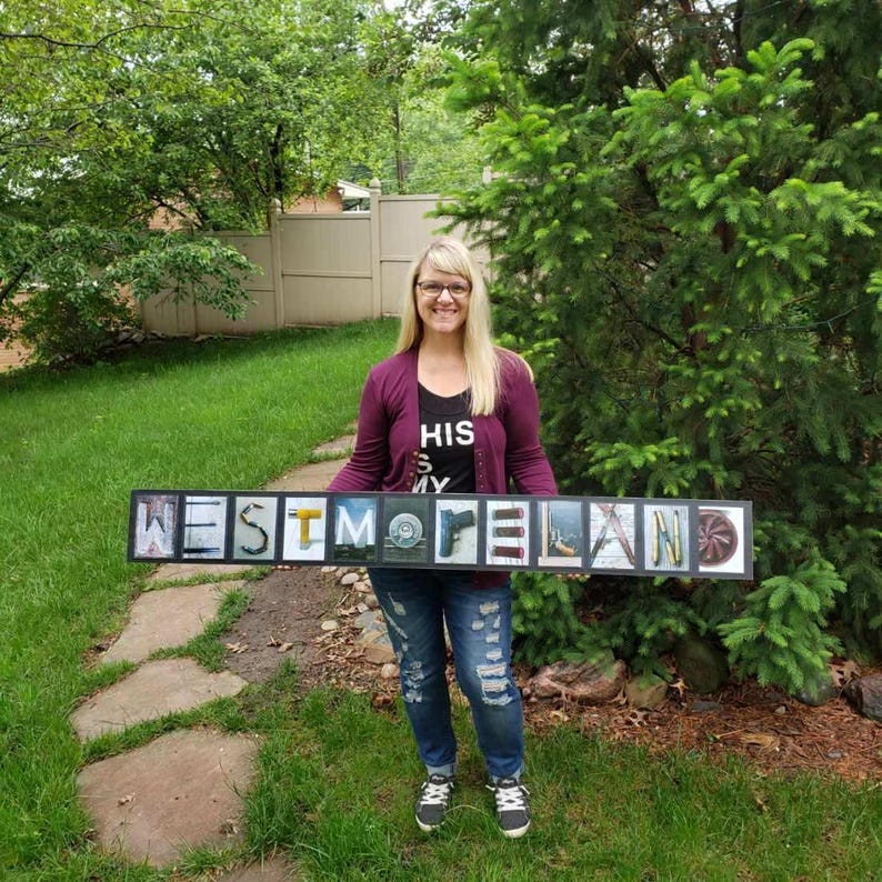Puede incluir: Una mujer sostiene un letrero enmarcado con la palabra "WESTMORELAND" en letras individuales. El marco es negro, las letras blancas y las letras contienen im&aacute;genes. La mujer lleva una camiseta negra, una rebeca burdeos y vaqueros azules.