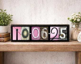 May include: A black framed wooden sign with the numbers "10 06 25" displayed. The numbers are made of various materials and set against a pink and wood background. The sign is on a wooden shelf with plants and flowers in the background.