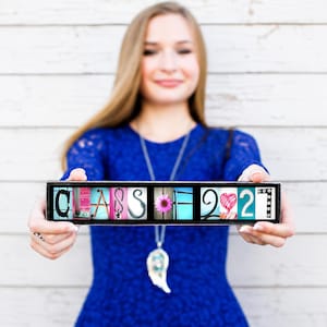 Puede incluir: Un letrero rectangular con el texto CLASS OF 2027, donde cada letra y n&uacute;mero est&aacute; representado por una imagen fotogr&aacute;fica &uacute;nica. Las letras se muestran en un marco negro, exhibiendo diversas texturas y colores, incluyendo una flor rosa.