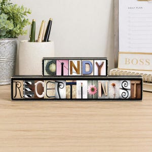 May include: A two-tiered black wooden sign displaying the name CINDY and the title RECEPTIONIST. Each letter is formed by a unique photograph of objects resembling that letter, including a pink flower, a pencil, and various metal shapes.