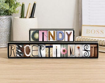 May include: A two-tiered black wooden sign featuring the name CINDY on the top row and the word RECEPTIONIST on the bottom row. Each letter is represented by a unique photograph of an object shaped like that letter, arranged in a frame.