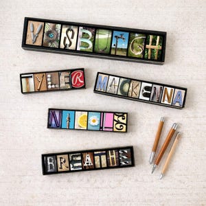 May include: Several personalized name signs and a sign that says "Breathe" are displayed. Each sign features letters with unique backgrounds, including images and textures. Three wooden pens with silver tips are also visible.
