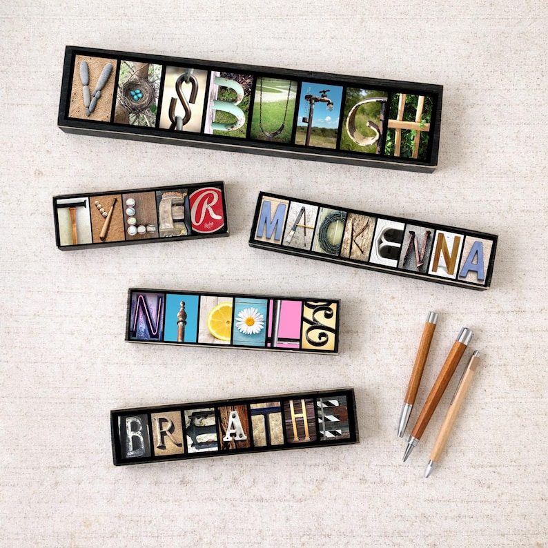 May include: Several personalized name signs and a sign that says "Breathe" are displayed. Each sign features letters with unique backgrounds, including images and textures. Three wooden pens with silver tips are also visible.