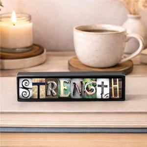 May include: A decorative wooden block with the word "STRENGTH" spelled out in various textured letters. The block is black and sits on a stack of books. A lit candle and a mug of coffee are in the background.