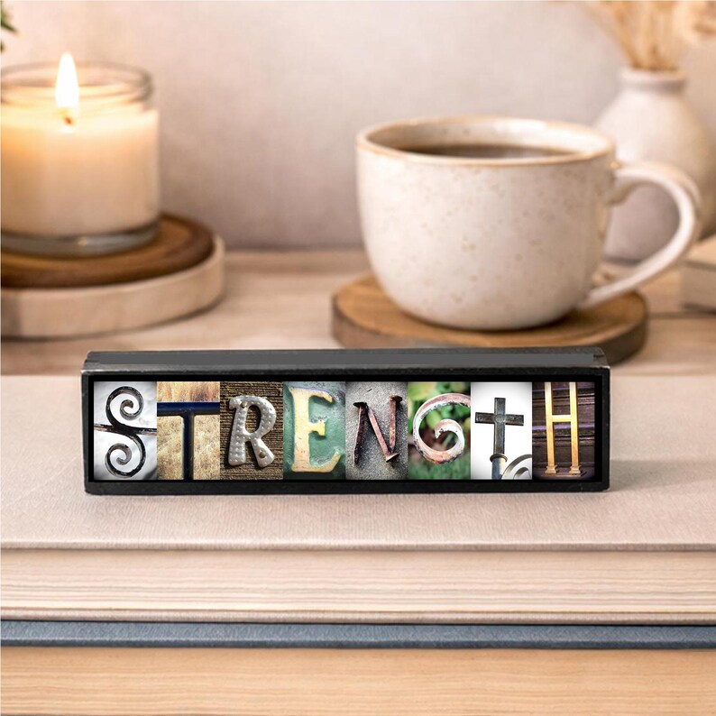 May include: A decorative wooden block with the word "STRENGTH" spelled out in various textured letters. The block is black and sits on a stack of books. A lit candle and a mug of coffee are in the background.