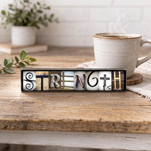 May include: A wooden block sign with the word "STRENGTH" spelled out in various fonts and textures. The letters are set against a black background. A coffee mug and plant are in the background, suggesting a cozy, inspirational theme.