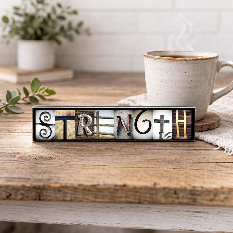 May include: A wooden block sign with the word "STRENGTH" spelled out in various fonts and textures. The letters are set against a black background. A coffee mug and plant are in the background, suggesting a cozy, inspirational theme.