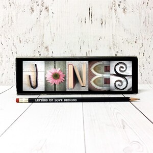 May include: A black rectangular block displaying the name "JONES" with each letter represented by a different image: a metal hook, a pink flower, books, a church window, and a decorative scroll. A pencil with the text "LETTERS OF LOVE DESIGNS" lies below.