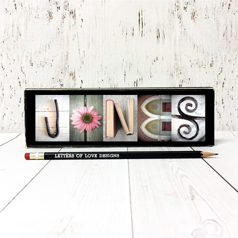 May include: A black rectangular block displaying the name "JONES" with each letter represented by a different image: a metal hook, a pink flower, books, a church window, and a decorative scroll. A pencil with the text "LETTERS OF LOVE DESIGNS" lies below.