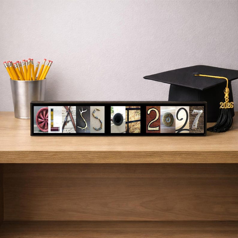 Puede incluir: Un letrero rectangular enmarcado que muestra la palabra CLASS 2027, donde cada letra y n&uacute;mero est&aacute; formado por una fotograf&iacute;a de un objeto. El letrero est&aacute; junto a un portal&aacute;pices plateado con l&aacute;pices amarillos y un birrete negro.