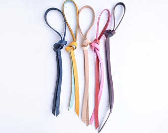 May include: Five leather tassels in various colors: navy blue, yellow, light blue, tan, pink, and burgundy. Each tassel has a knotted loop at the top and a split, fringed bottom. The tassels are arranged against a white background.