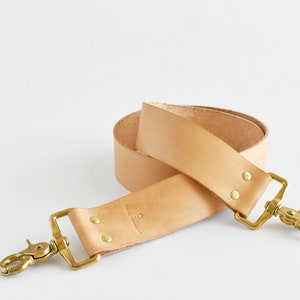 May include: A natural-toned leather strap with brass-colored metal hardware. The strap has two brass clasps and is secured with rivets. The text "China Merchant" is stamped on the leather. The strap is rolled up.