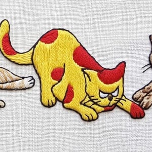May include: Embroidered cat design with yellow and red accents. The cat is in a playful pose, with a grumpy expression. The embroidery is on a white fabric background. The design features a cartoon cat.