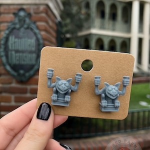 May include: A pair of gray gargoyle earrings displayed on a brown earring card. The earrings are shaped like cartoon gargoyles with their arms raised. The background features a sign that says "Haunted Mansion".