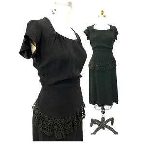 May include: A black dress featuring short sleeves, a square neckline with subtle gathering, and a fitted bodice. The waistline is accented with a decorative black lace trim that extends into a peplum-style detail over the skirt portion.