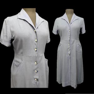 May include: A light blue button-down dress with short sleeves, a collared neckline, and two side pockets. The dress features a row of decorative buttons down the front, each with a small floral pattern, and a textured fabric finish throughout.