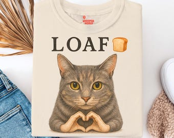 May include: Cream-colored t-shirt with the word "LOAF" above a cat illustration. The cat is gray and brown with yellow eyes, forming a heart shape with its paws. A small bread graphic is next to the word "LOAF".