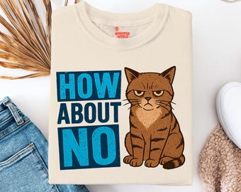 Op de afbeelding: Crèmekleurig T-shirt met een illustratie van een chagrijnige bruine kat en de tekst "HOW ABOUT NO" in blauw. Het shirt is opgevouwen, met een blauwe spijkerbroek en witte schoenen op de achtergrond. Het shirt heeft een klein logo aan de bovenkant.