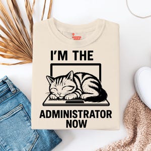 以下が含まれることがあります： 「I'M THE ADMINISTRATOR NOW」の文字と、ノートパソコンで寝ている猫のグラフィックがプリントされたクリーム色のTシャツ。猫は白と黒。Tシャツは丸首で半袖です。