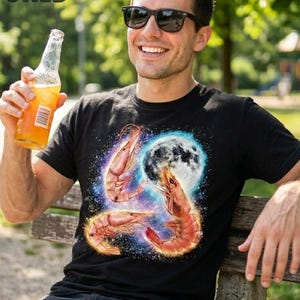May include: A black t-shirt featuring a graphic print of three orange shrimp surrounding a glowing full moon against a dark, starry nebula background. The text "WHOOPSIE WORLD" appears in red, bold, rounded letters in the upper left corner.